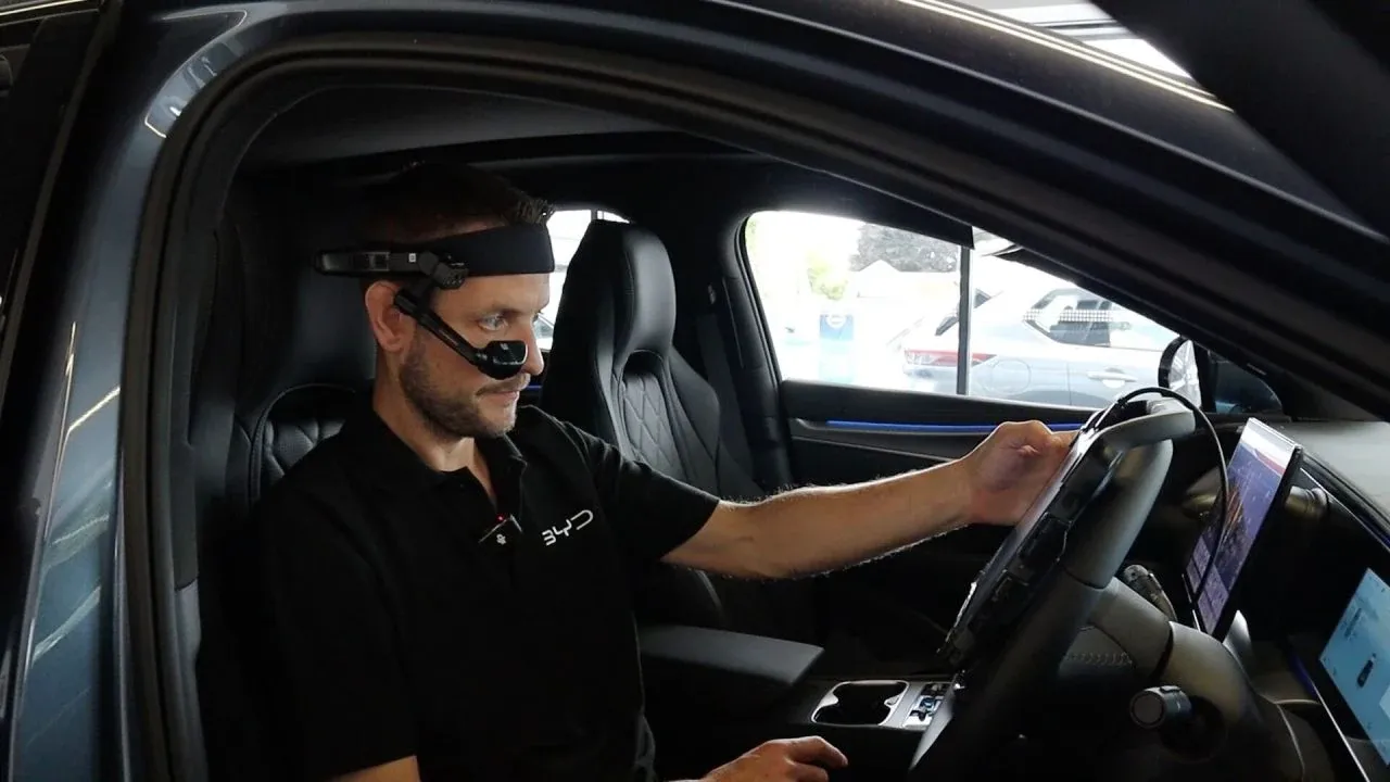 A BYD technician in the driver’s seat of a BYD car, wearing a RealWear head-mounted display running FieldPal, with one hand on the steering wheel